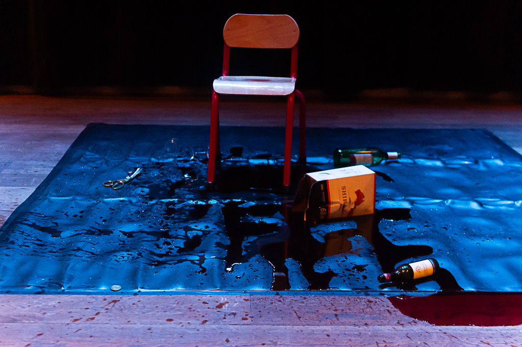 A chair on a tarpaulin splattered with wine, with some of the wine leaking off the edge onto a wood panelled floor.