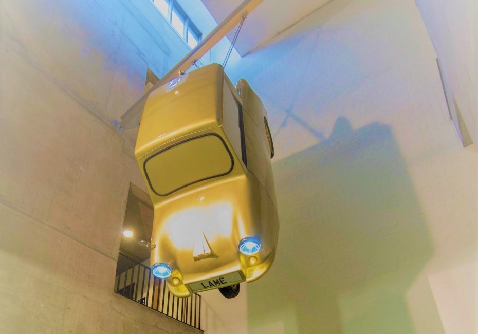 A gold 'Invacar' vehicle suspended from a beam in a high-ceilinged room. The numberplate on the car read 'GOLD LAMÉ'