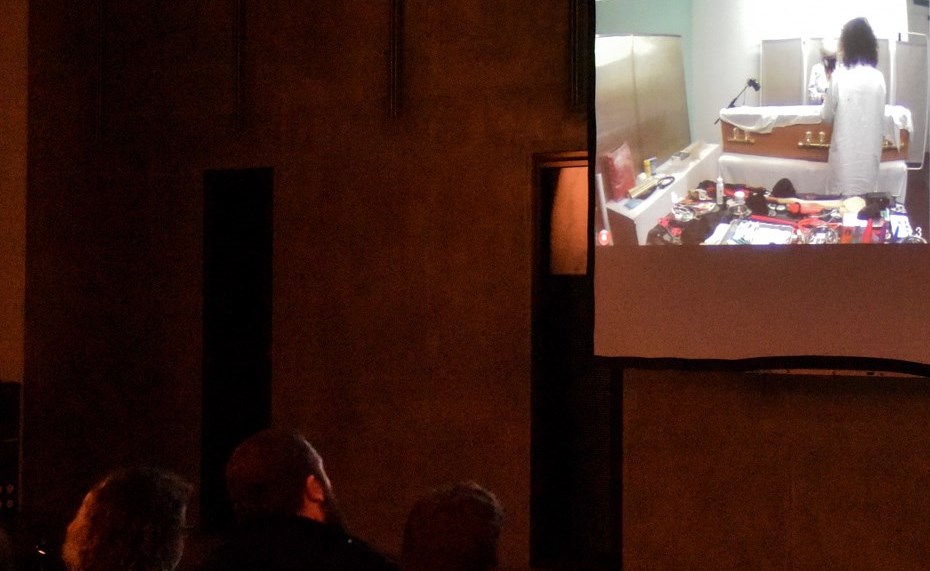 Peopl in a dark room watching a screen which displays footage of a coffin.