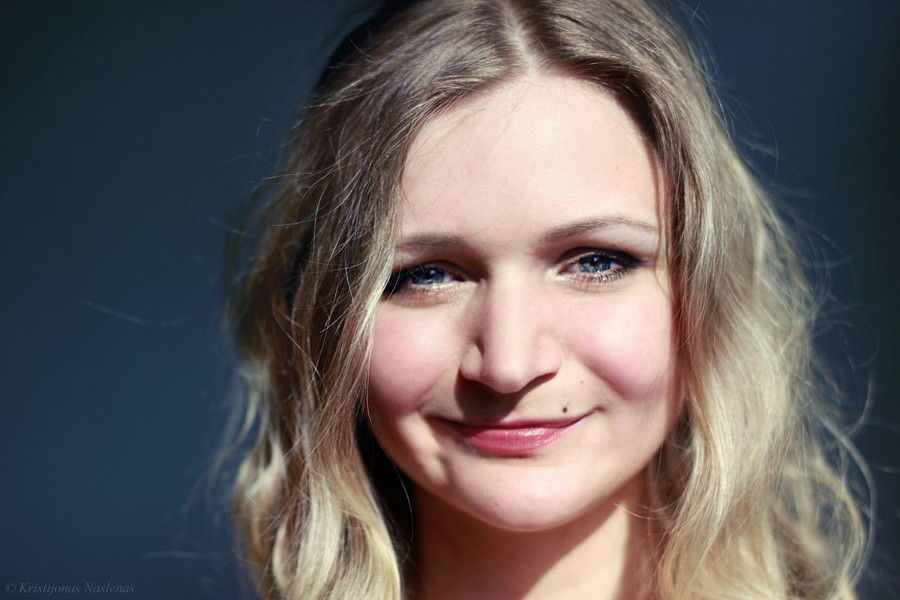 A blonde woman with blue eyes smiling against a dark blue backdrop.