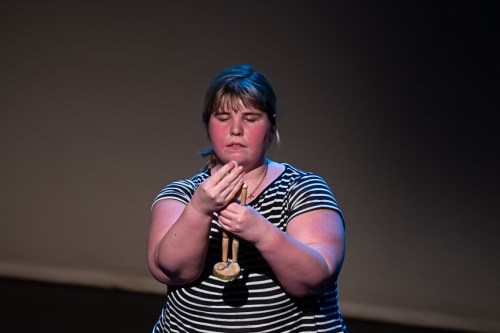 Bethany Murray, a performer wearing a black and white striped top, stands onstage, transfixed by a small wooden mannequin she holds before her.