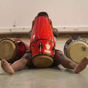 A black man sitting on the floor is obscured by three drums