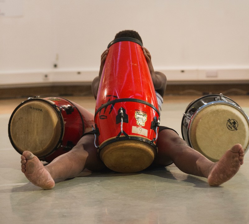 A black man sitting on the floor is obscured by three drums