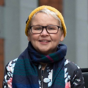 Nickie smiles into camera - she is outdoors wearing a woolly pullover and scarf.. Nickie is a wheelchair user.